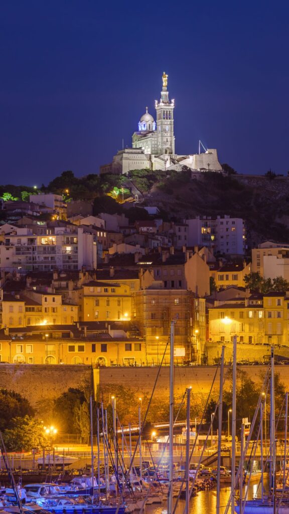 Notre-Dame de la Garde illuminée, vue de nuit sur le Vieux-Port de Marseille, avec les toits de la ville en contrebas.